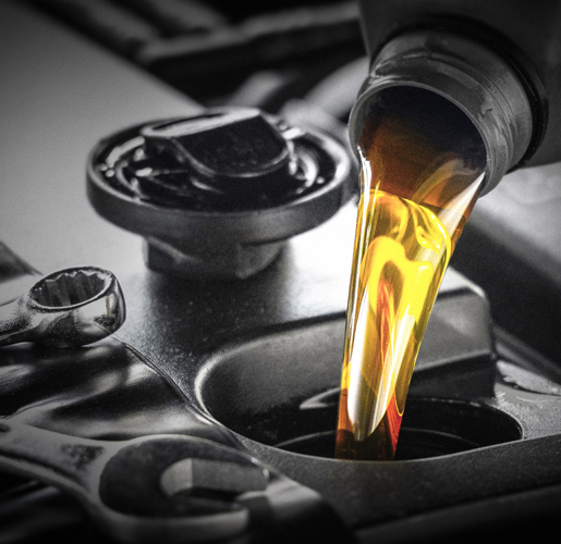 The image shows golden motor oil being poured from a container into an engine oil fill opening, with metal tools and engine components visible in the foreground.