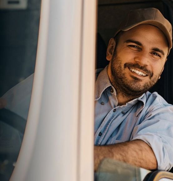 The image shows a truck driver seated inside a truck cab, viewed through the open side window, with the steering wheel and side mirror visible.