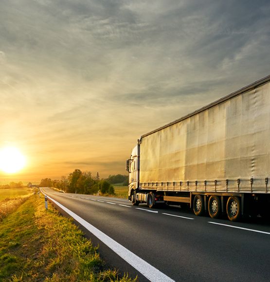 The image shows a large semi-truck with a covered trailer driving along a rural road at sunset, with open fields on both sides and the sun low on the horizon.